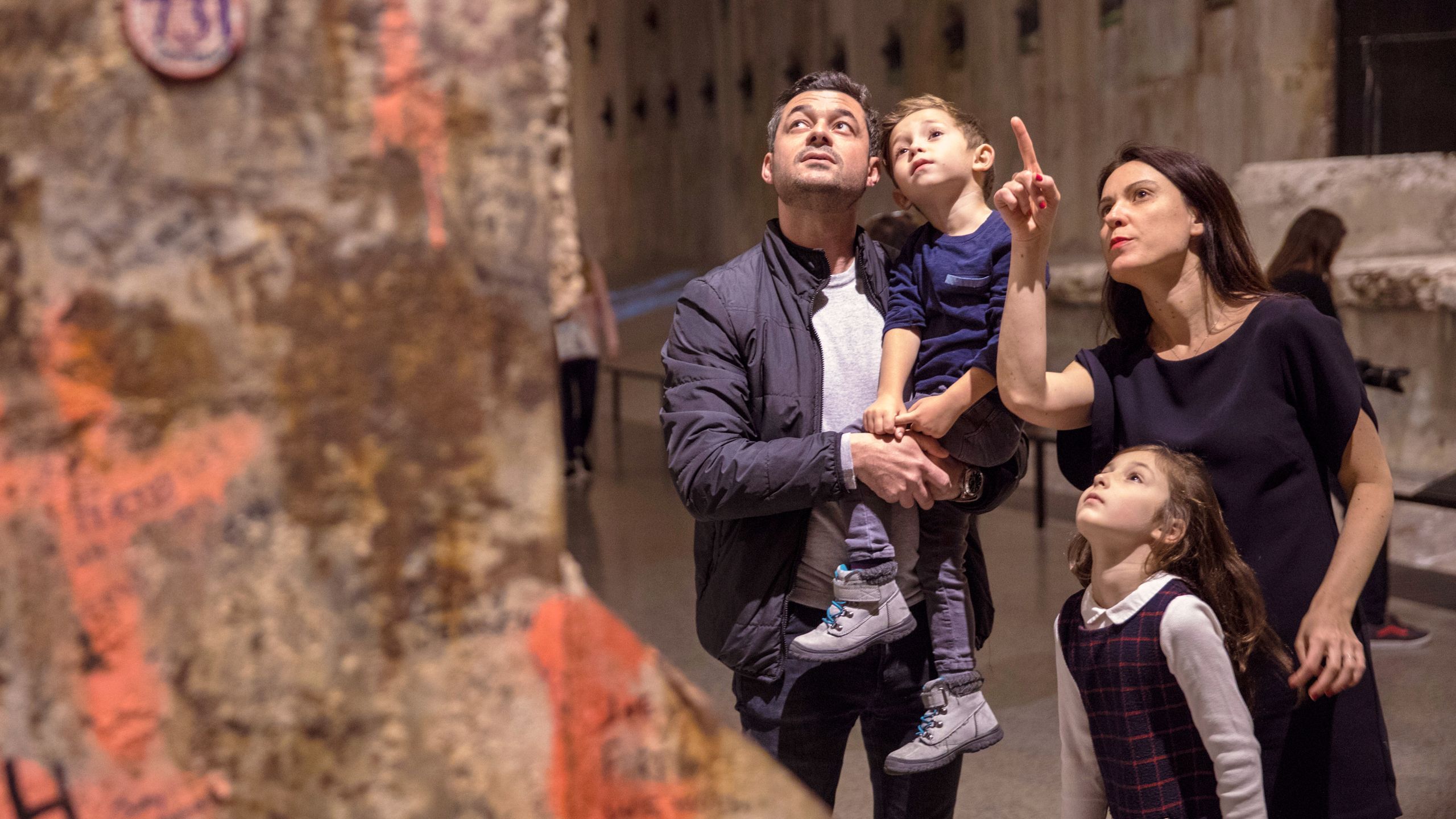 A man and woman stand with two a young boy and girl at the Museum. The man is holding the boy as the woman stands with the girl by her side. All four of them are looking up at the Last Column as the woman points up at it.