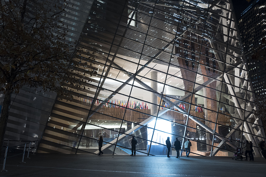 A night view shows several people walking by the lit up 9/11 Memorial Museum. Several darkened trees are in the foreground.