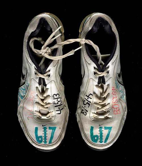 Two sneakers left at a spontaneous memorial in Copley Square in Boston are displayed on a black surface. The sneakers have the date of the Boston Marathon Bombing—April 15, 2013—written on them in marker.