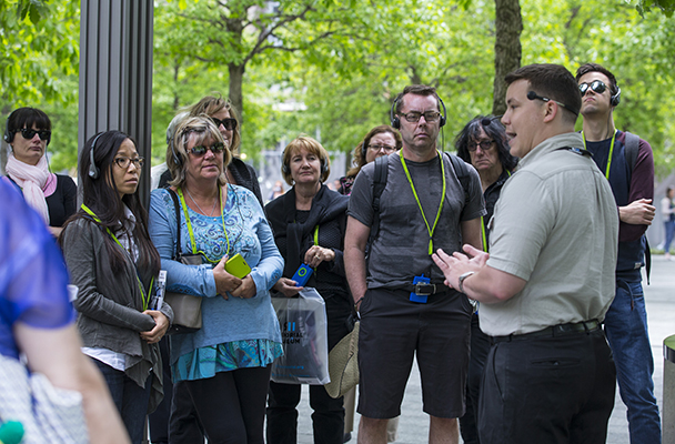 A 9/11 Memorial tour guide tells visitors about the Memorial. About a dozen visitors watch on as he speaks to them.