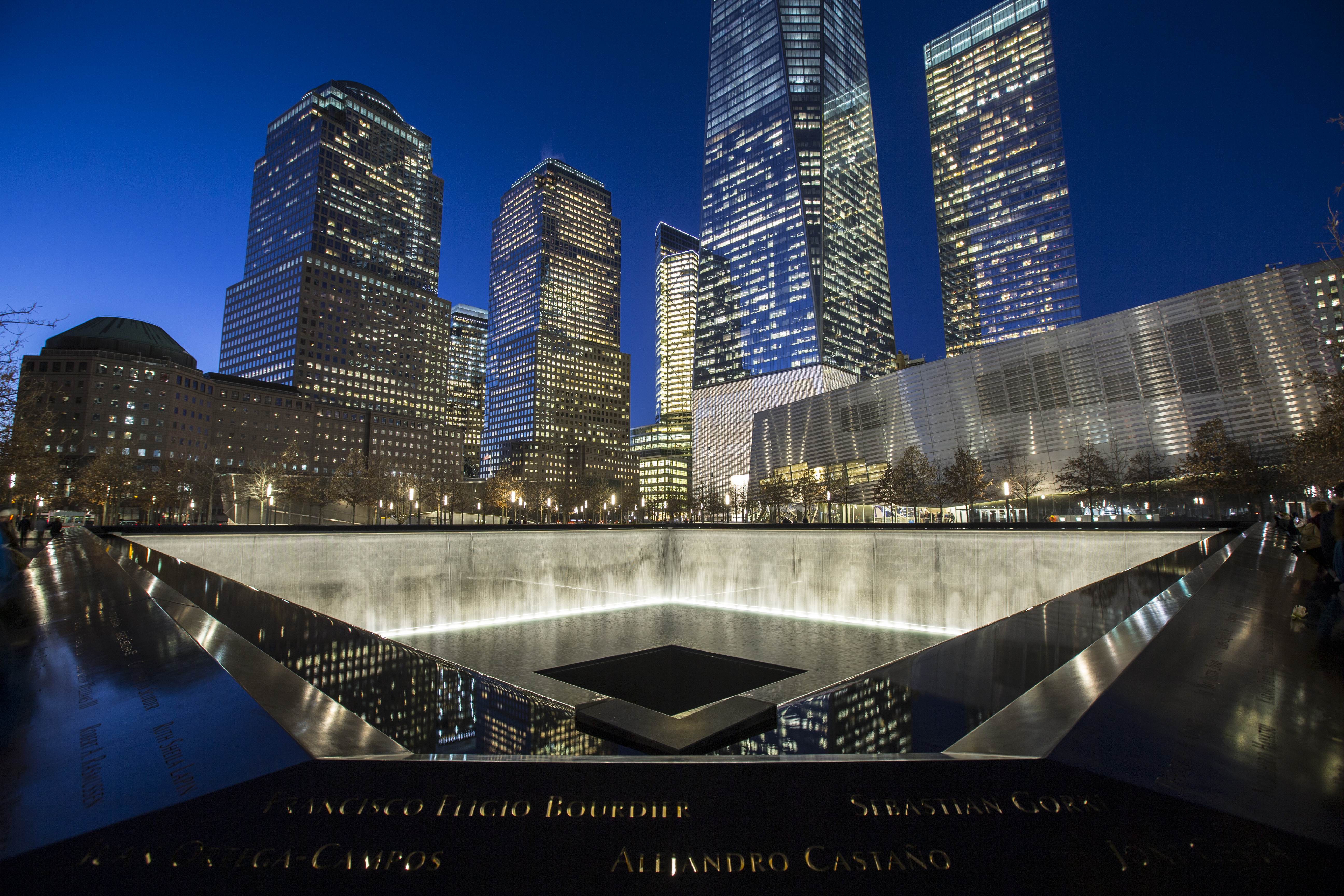 One World Trade Center towers over the south pool of the 9/11 Memorial on a clear night. Water cascades down the illuminated reflecting pool.