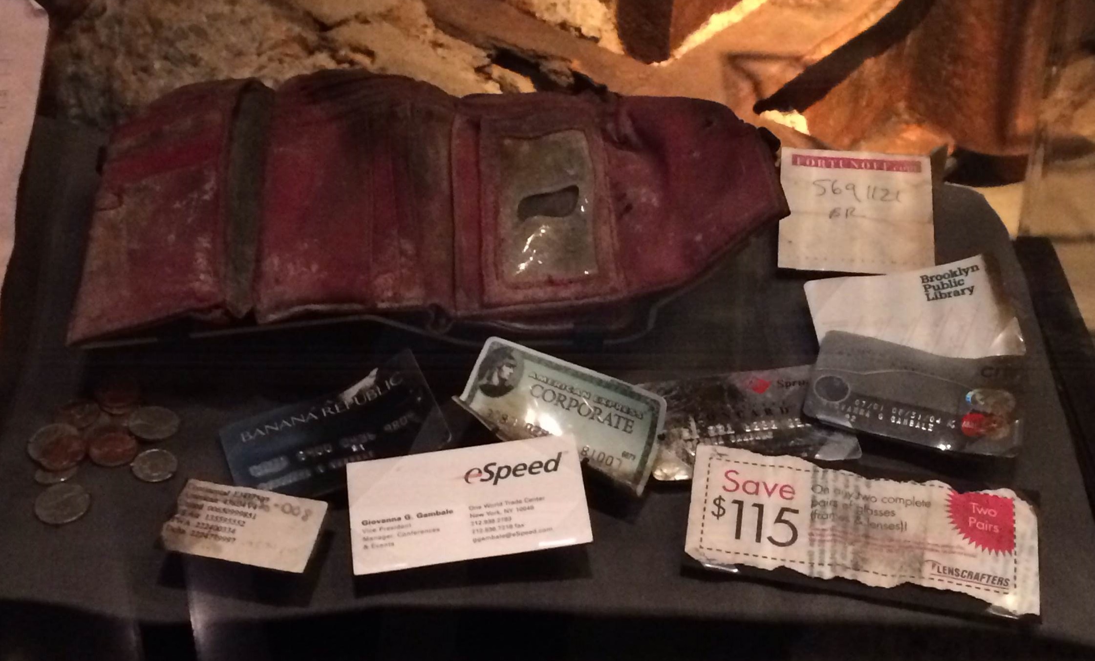 A wallet belonging to Giovanna Gambale is displayed at the Museum. The red wallet is heavily damaged and stained with dirt. The contents of the wallet, including credit cards, coupons, note paper, and coins have been laid out in front of it.