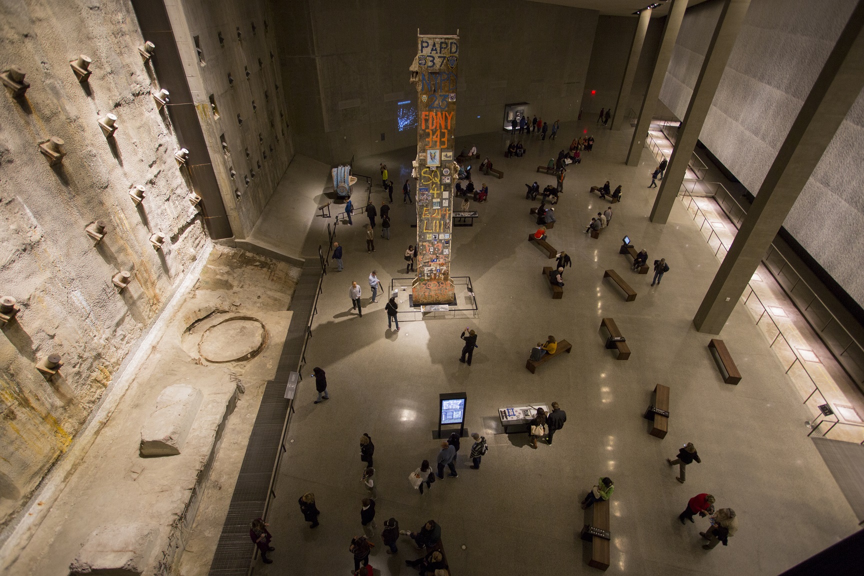 A view over Foundation Hall at the Museum shows the slurry wall, Last Column, and dozens of visitors sitting on benches, looking at artifacts, or interacting with touchscreens.