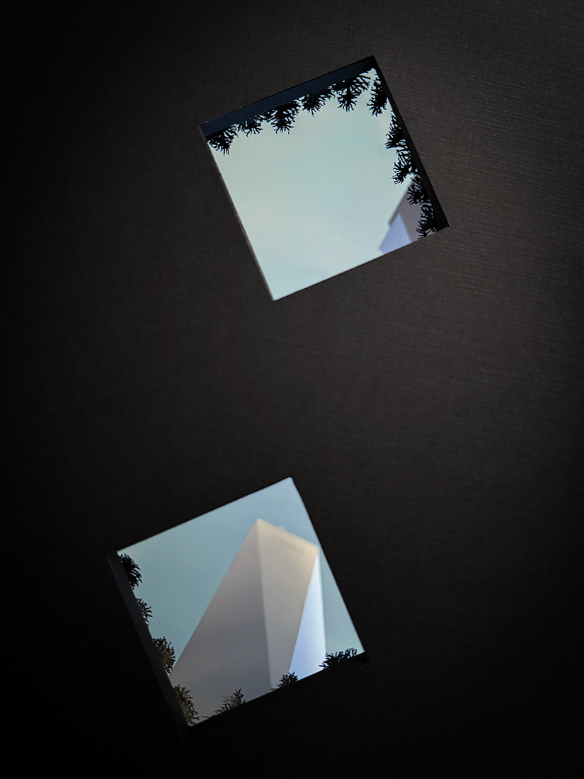 Paper tunnel-book sculpture in layered black frames, viewed from inside two square voids representing the World Trade Center memorial pools; a white paper tower and cluster of buildings rise beyond a line of dark green trees.