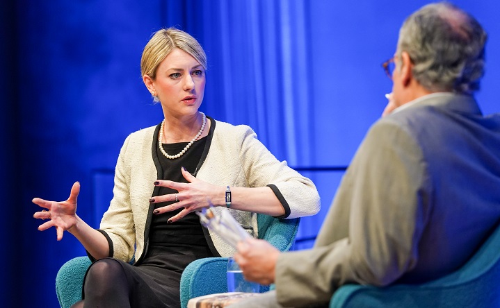 Joana Cook, public program speaker, gestures on a blue auditorium stage in conversation with Cliff Chanin, whose back is to the camera.