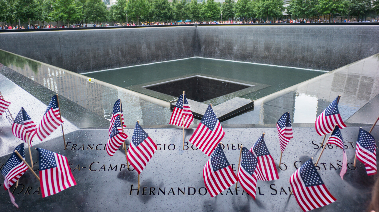 American flags are placed in the names of the 9/11 Memorial parapets. The center of the Memorial pool is visible behind them.