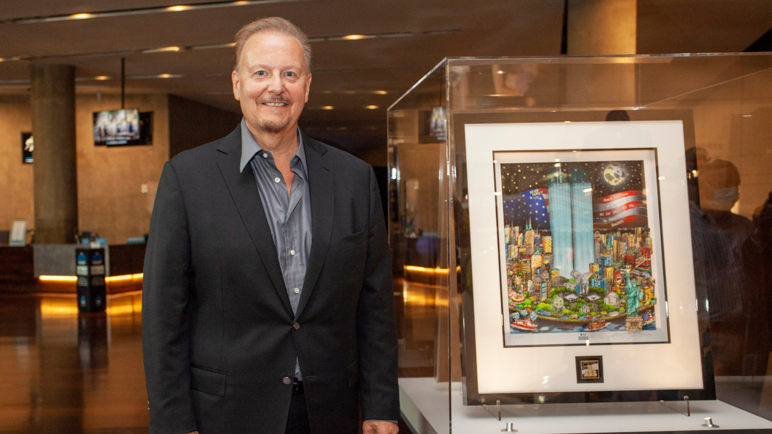 A bearded man in a dark jacket and gray shirt stands to the left of a display case containing his pop art painting of the Twin Towers