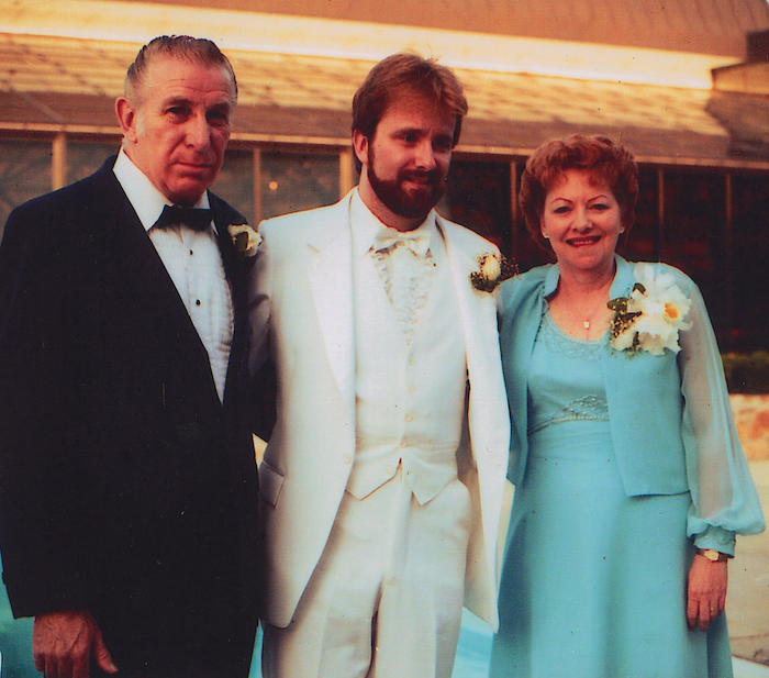 A bearded man in a white tuxedo stands between his father, on the left, in a black and white tuxedo, and his mother, on the right, in a turquoise dress