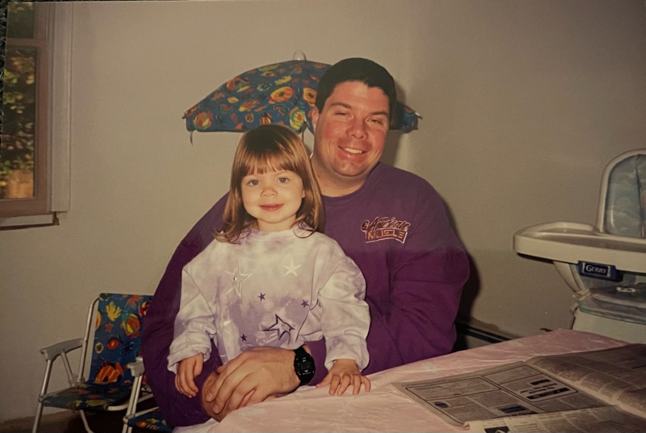 A young, blonde girl in a top with purple stars sits on her father's lap - he is wearing a burgundy top and has dark hair.