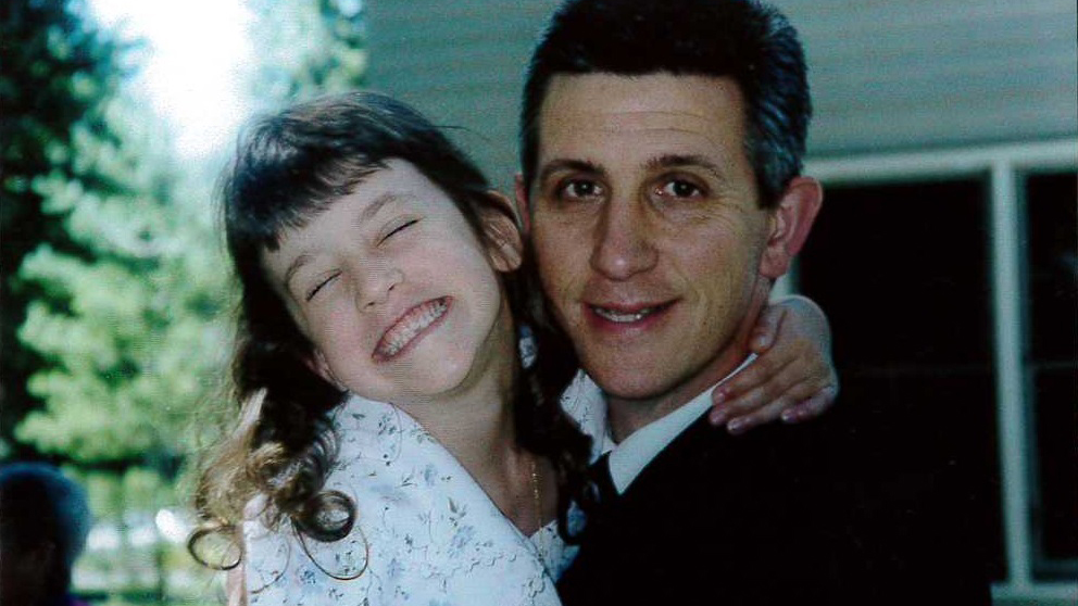 A young girl embraces her father, smiling so broadly that her eyes are closed.