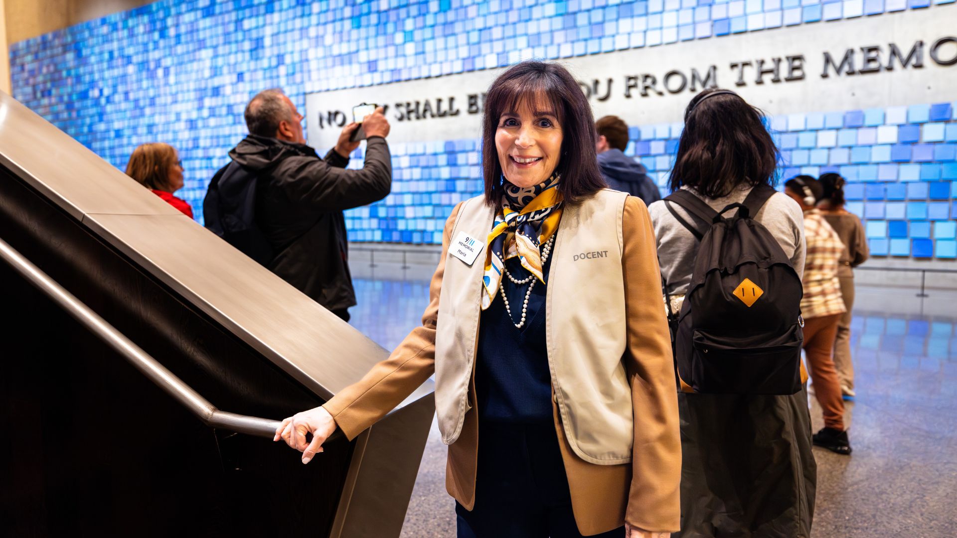 A docent in a beige vest stands at the foot of a staircase with a blue tiled art installation behind her
