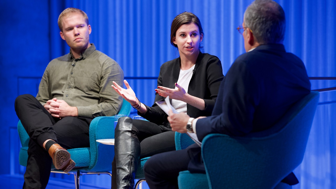 Two men and a woman sit on a blue-lit auditorium stage. The woman in the center gestures with her hands and addresses the moderator, whose back is to the camera.