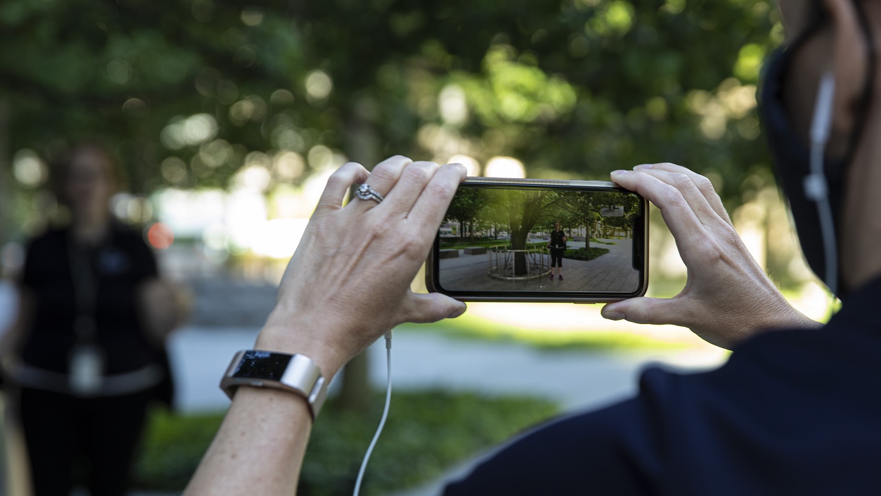 A close-up photo shows a smartphone recording a video of a woman speaking on the 9/11 Memorial plaza.