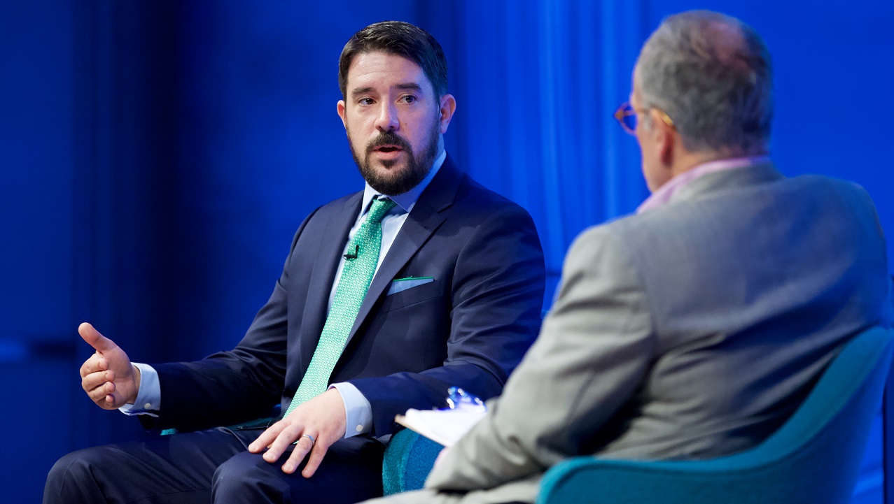 Two men sit on a blue-lit auditorium stage. One gestures with his hands.