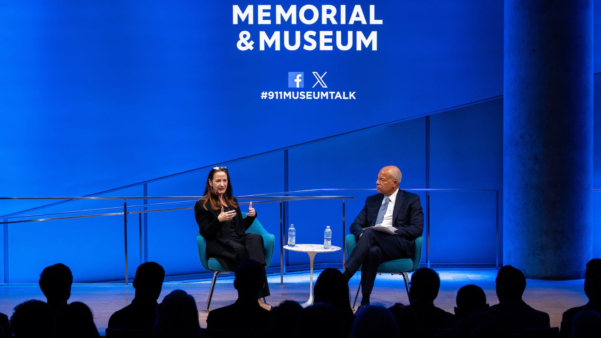 A woman (left) and a man sit on stage against a blue backdrop with a portion of the 9/11 Memorial & Museum logo visible. Both are wearing dark suits and the back of audience members' heads are visible in front.
