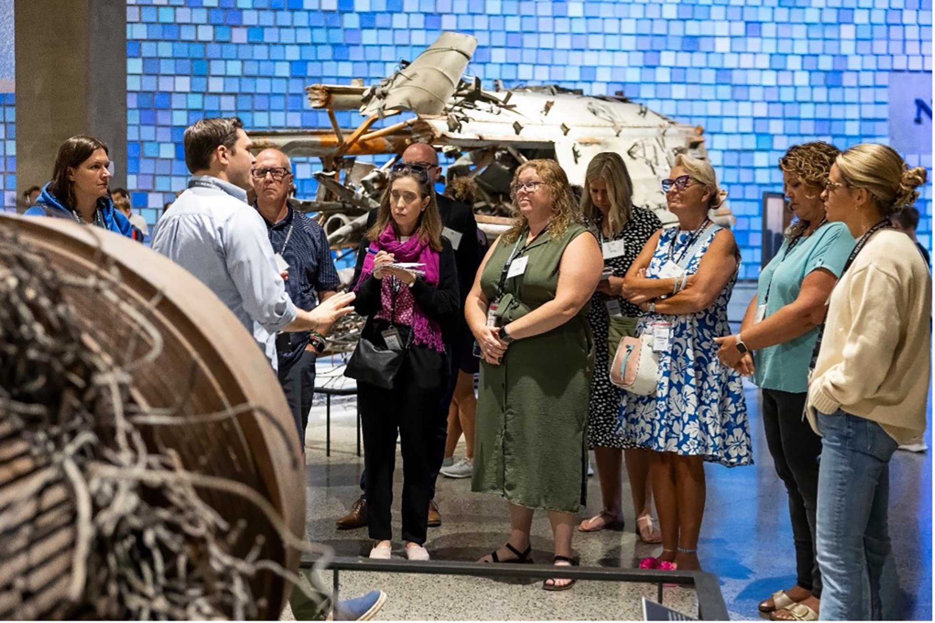 A man addresses a group of adults standing among large artifacts in the 9/11 Museum.