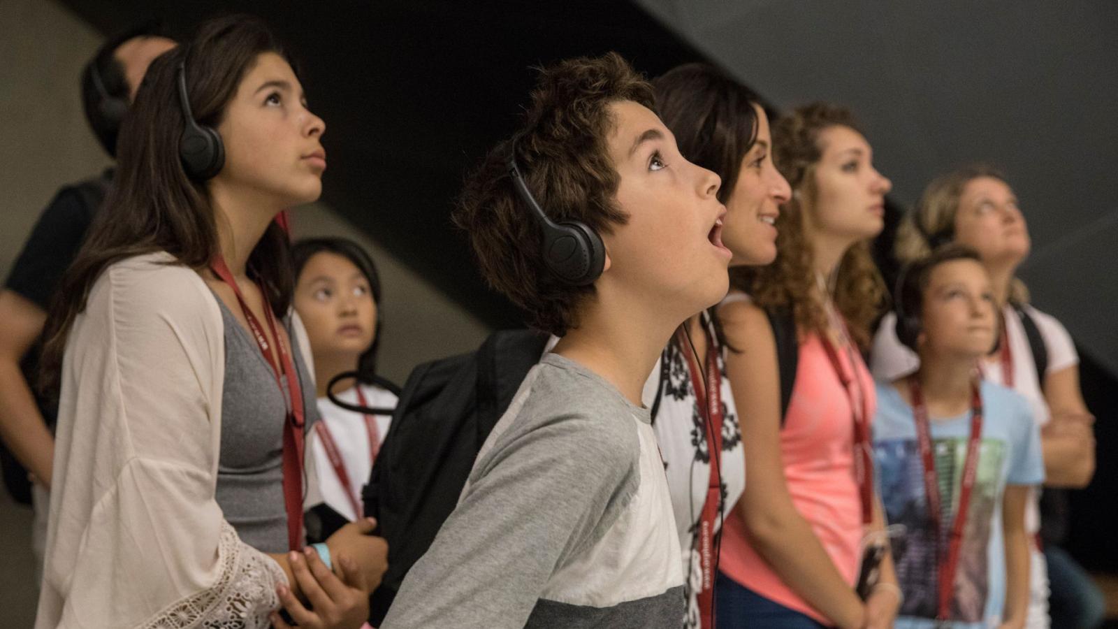 A small group of kids and adults wearing headphones look up in wonder