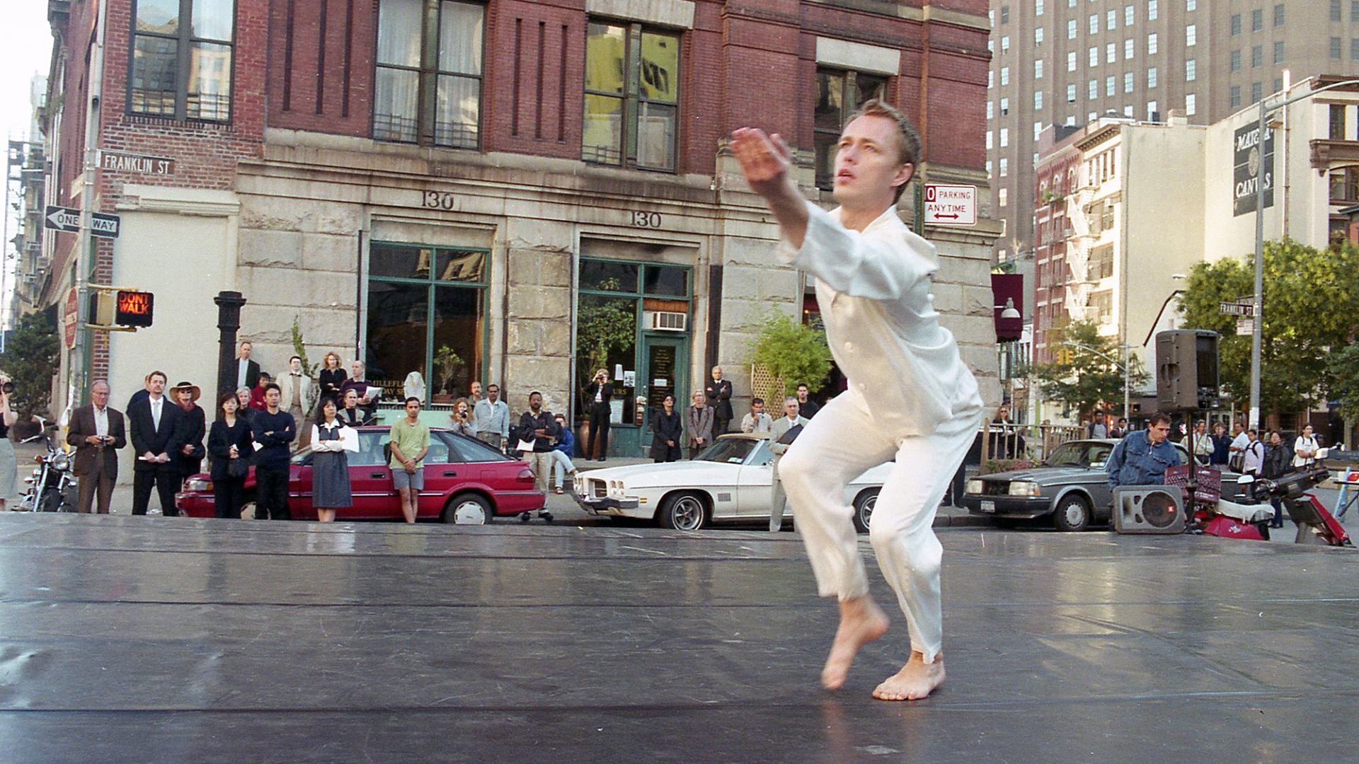A performer dressed in white dances in front of a small crowd and a brick building on Franklin Street.
