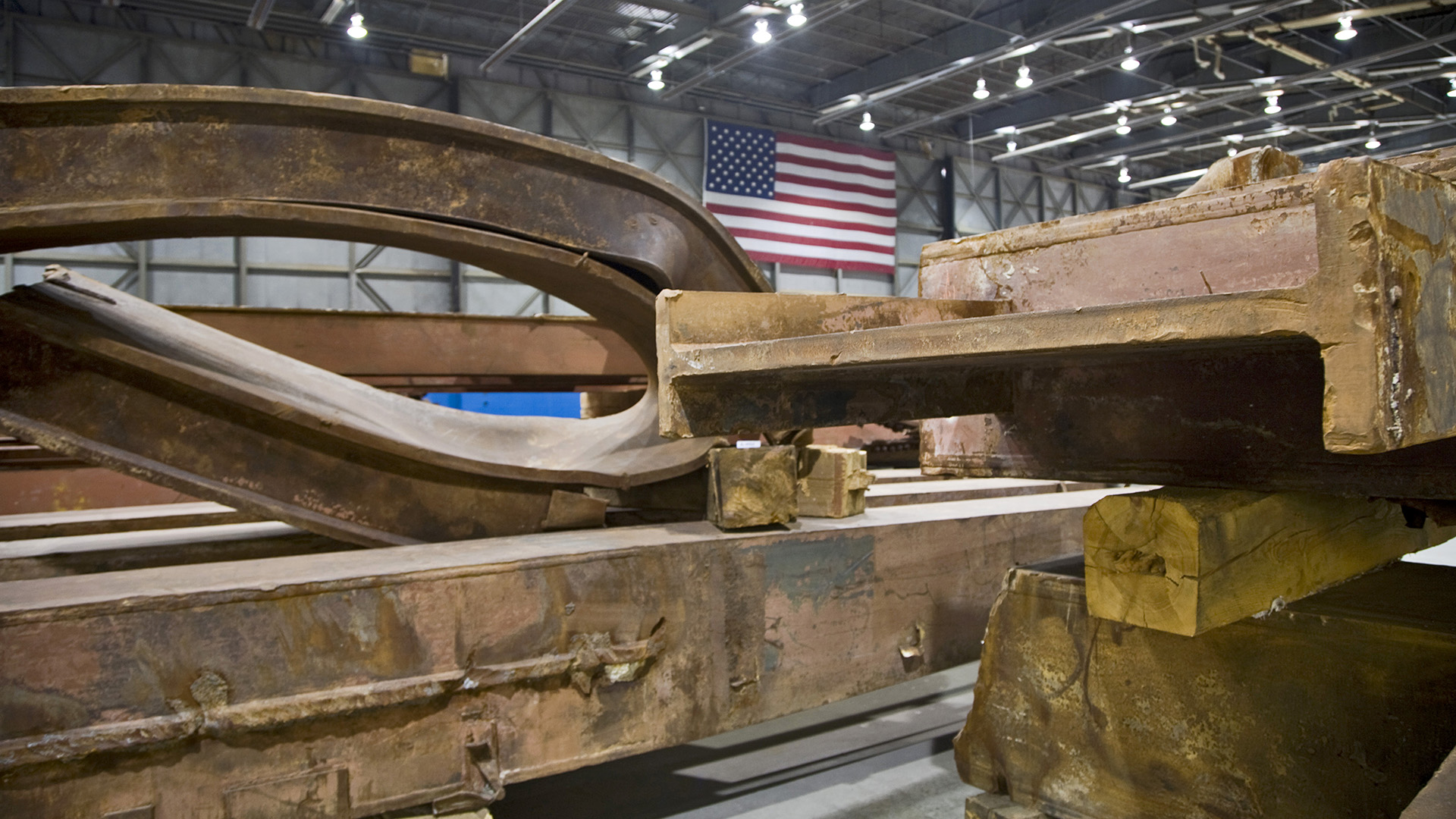 Rusted, warped metal beams and wood planks are piled near an American flag inside an airport hangar. 