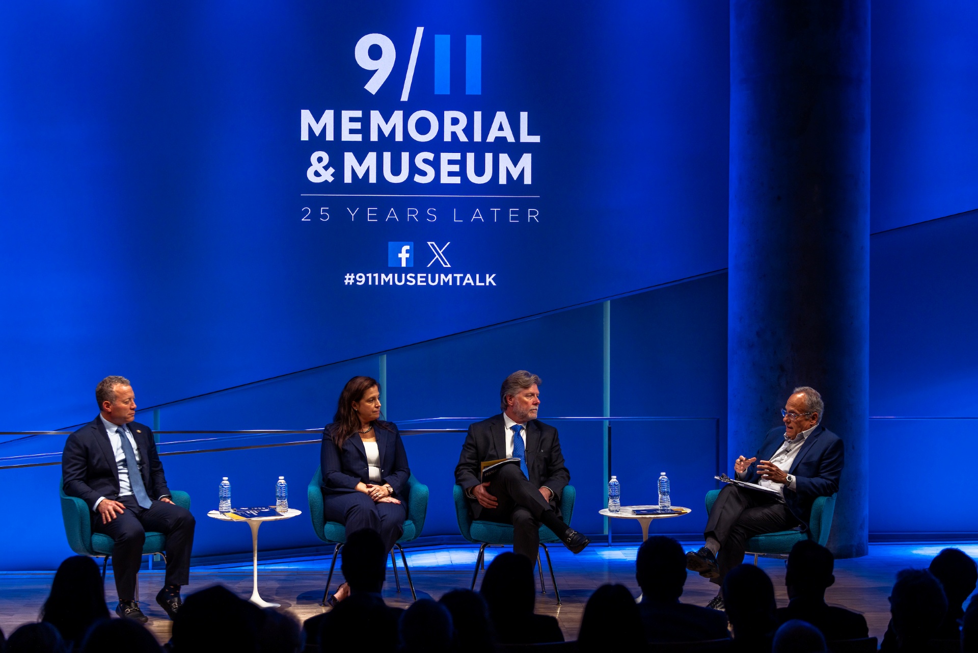 four people sit on stage in front of a blue background with the logo of the 9/11 Memorial & Museum behind them