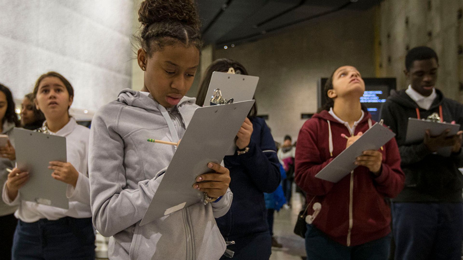Group of middle school students in different skin tones write on clipboards inside the museum, with aluminum cladding visible in the background.