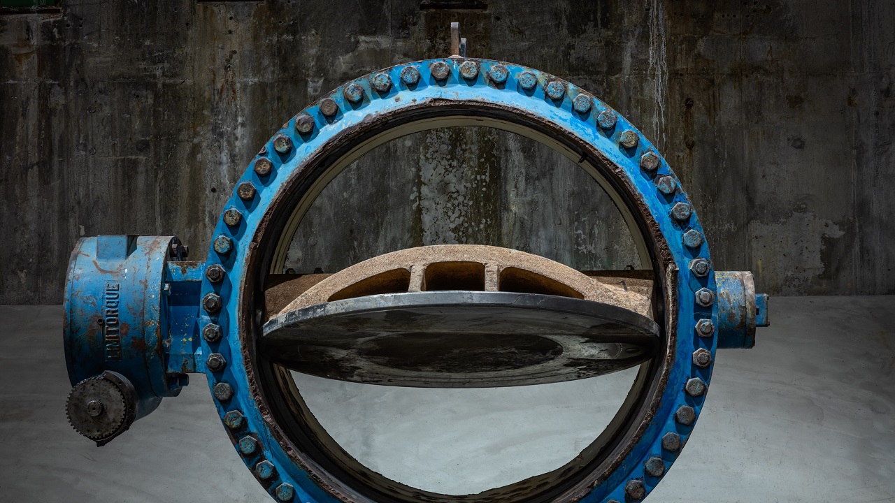 A blue, steel river water valve sits on a concrete floor. Screws line the border of the circular valve. It is in the open position, allowing the viewer to see through to the concrete wall behind it.