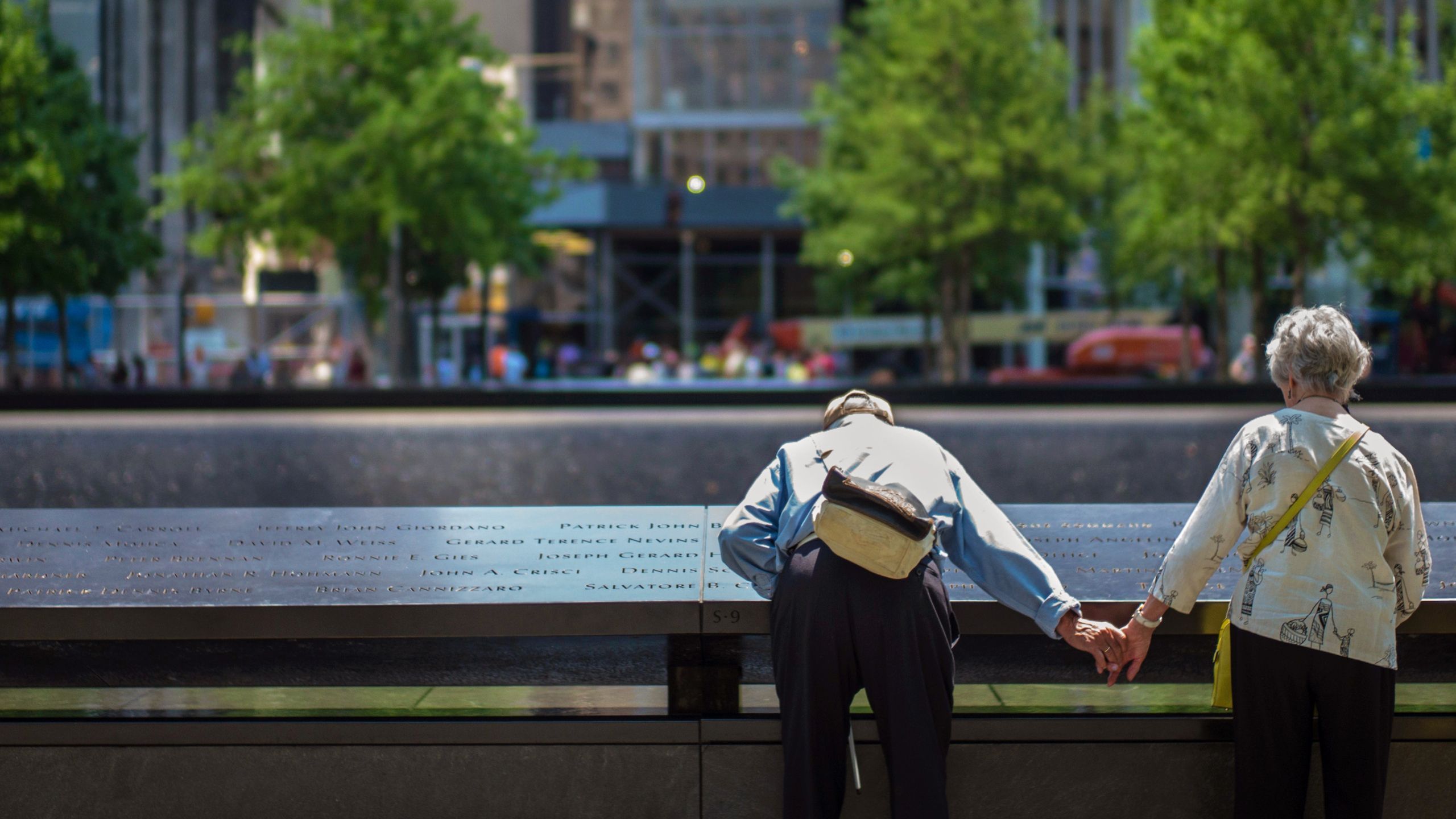 An elderly man holds hands with an elderly woman as he bends down to read names on the Memorial. Four green trees can be seen in the background, on the other side of the Memorial reflecting pool.