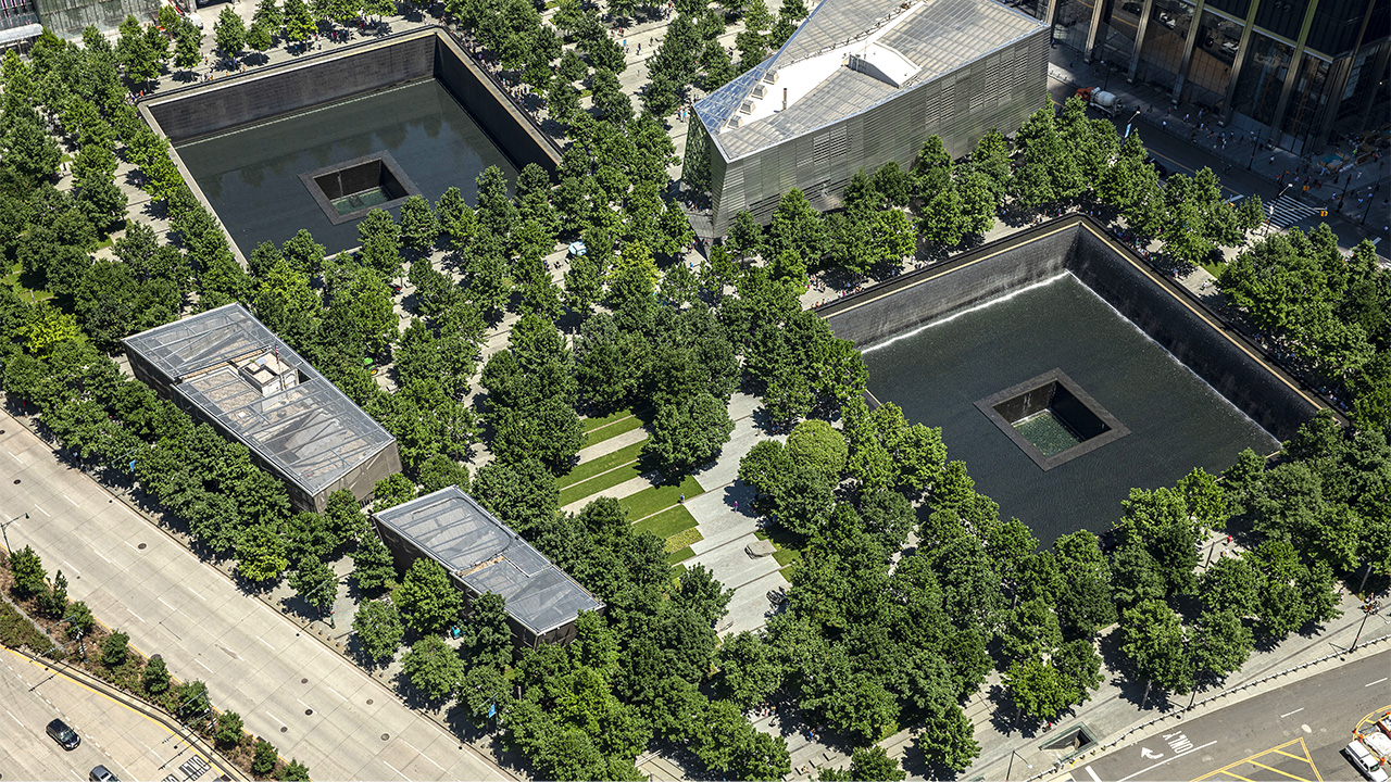 Memorial Plaza is seen from above in daylight. Dozens of oak trees fill the Plaza. These trees surround the two reflecting pools where the North and South towers once stood. Between the large, square pools is the Museum’s glass and steel pavilion.