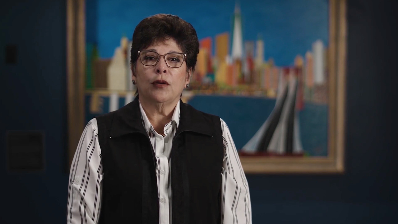 A woman with short black hair and glasses, wearing a blouse and black vest sits, talking, in front of a painting of downtown Manhattan.