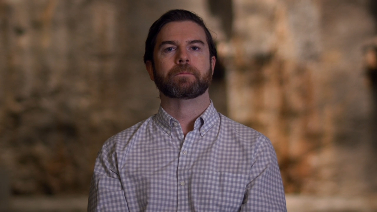 A bearded man wearing a button-up shirt sits and talks.