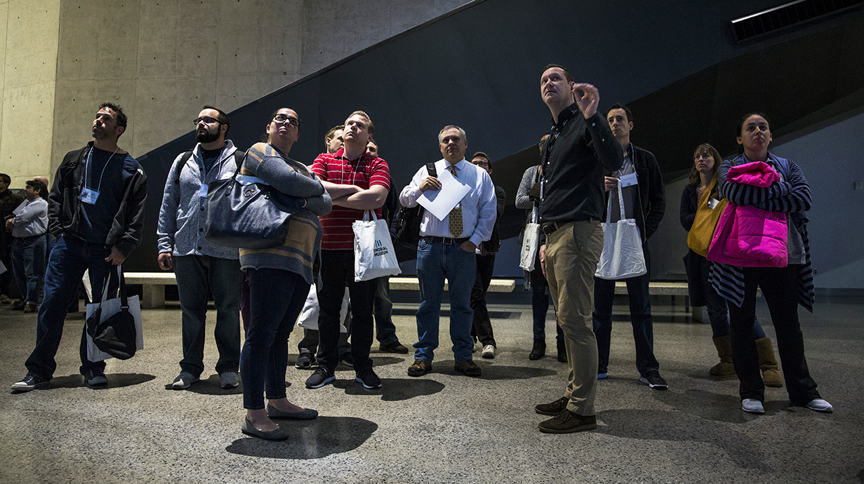 An instructor leads a teacher workshop at the Museum. He and a group of men and women are standing and facing forward as they observe something out of view.