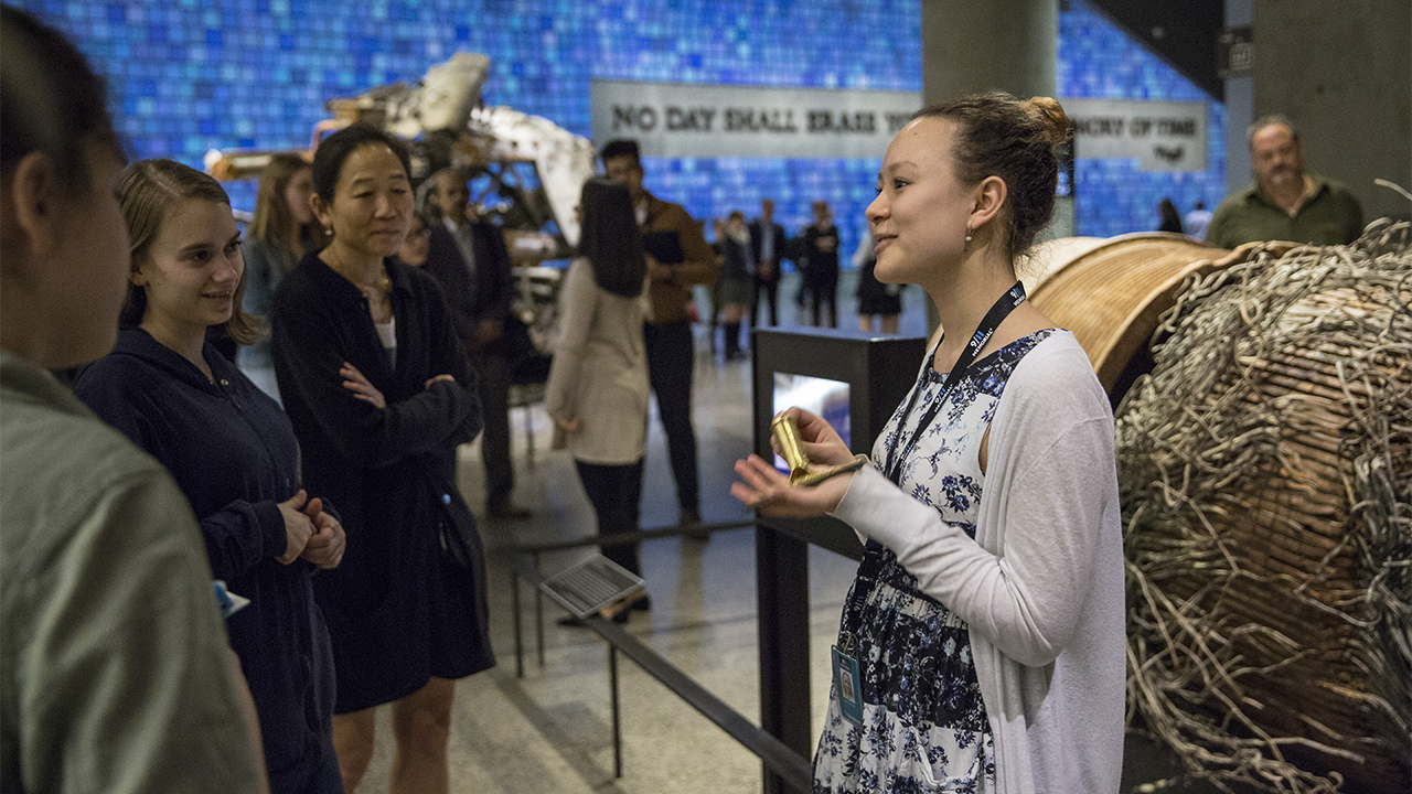 A young woman holds a gold-colored squeegee as she speaks to several women at the Museum. She stands beside a destroyed elevator motor from the Twin Towers.