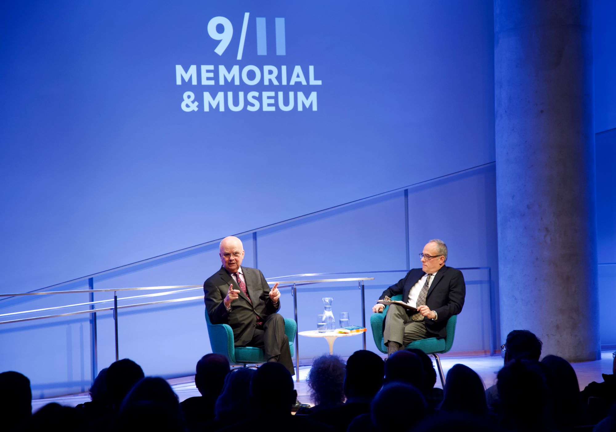 Former CIA director and retired U.S. Air Force Gen. Michael Hayden speaks onstage as part of the public program, General Michael Hayden on the War on Terror. Hayden gestures with both hands as he looks out at the audience, which is silhouetted by the lights of the stage. Clifford Chanin, the executive vice president and deputy director for museum programs, is seated next to him.