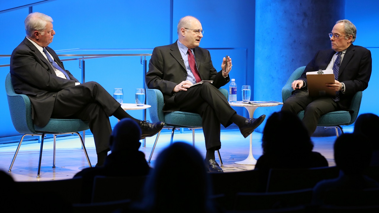 In this photograph, two participants in a public program sit on a blue-lit auditorium stage with legs crossed in suits and ties. One man in the center of the stage is speaking and gesturing toward the moderator, who sits holding a clipboard in his lap. On the other side of the man speaking sits the other public program participant, who sits with an expression of patient listening. Silhouetted in the foreground of the photograph are the heads of the audience members.