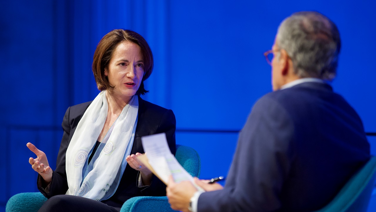 A woman in a gauzy scarf addresses a man in a suit, who holds a clipboard and is facing away from the camera.