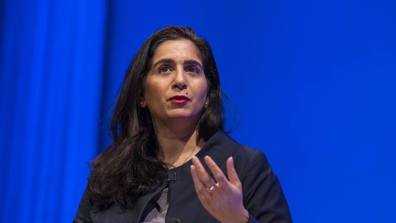 In this close-cropped photograph, a woman sits in front of a blue background and gestures with her left hand as she speaks.