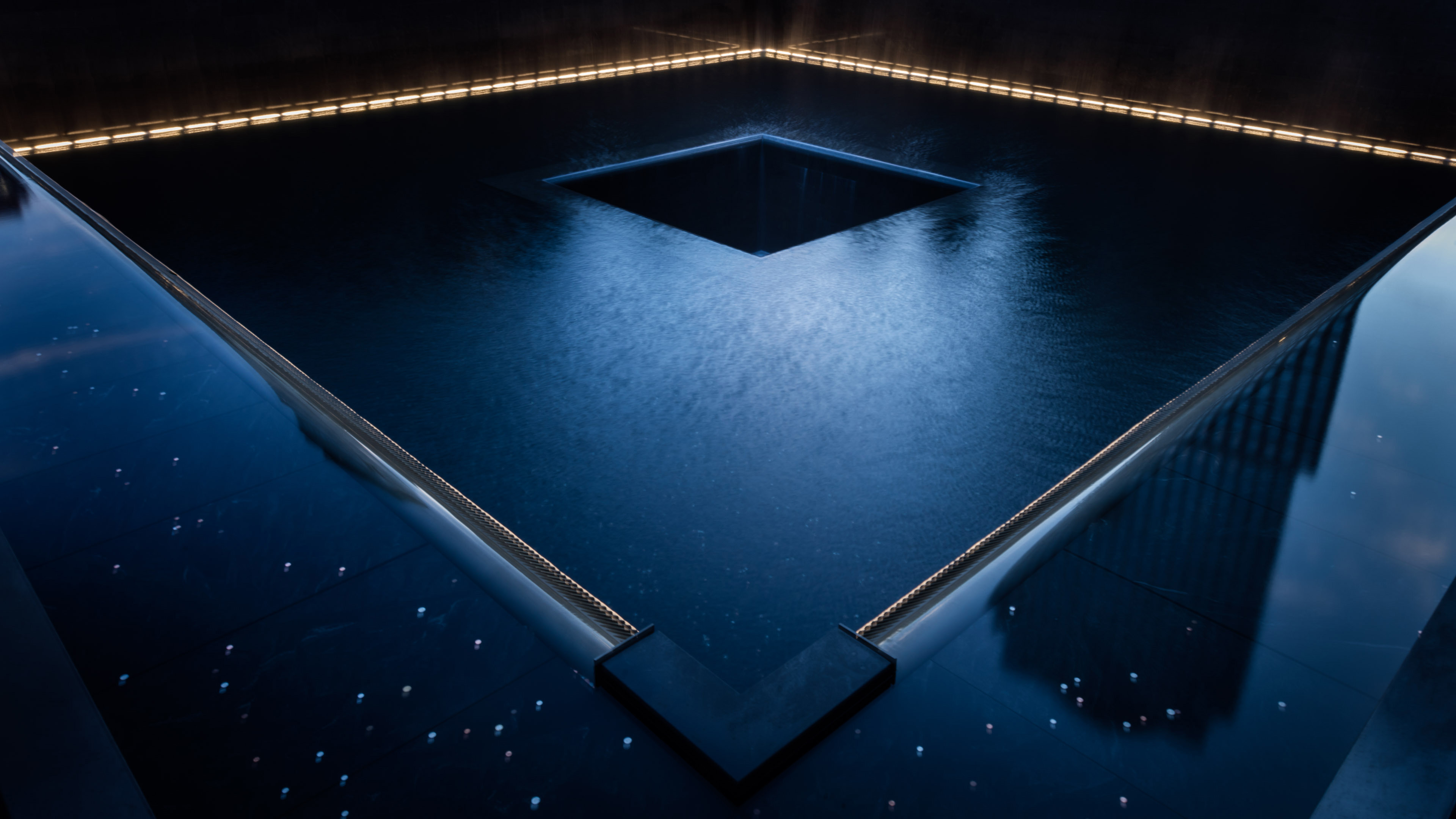A memorial pool is illuminated at night. Yellow lights are glow in the background as bluish light is reflected in the foreground.