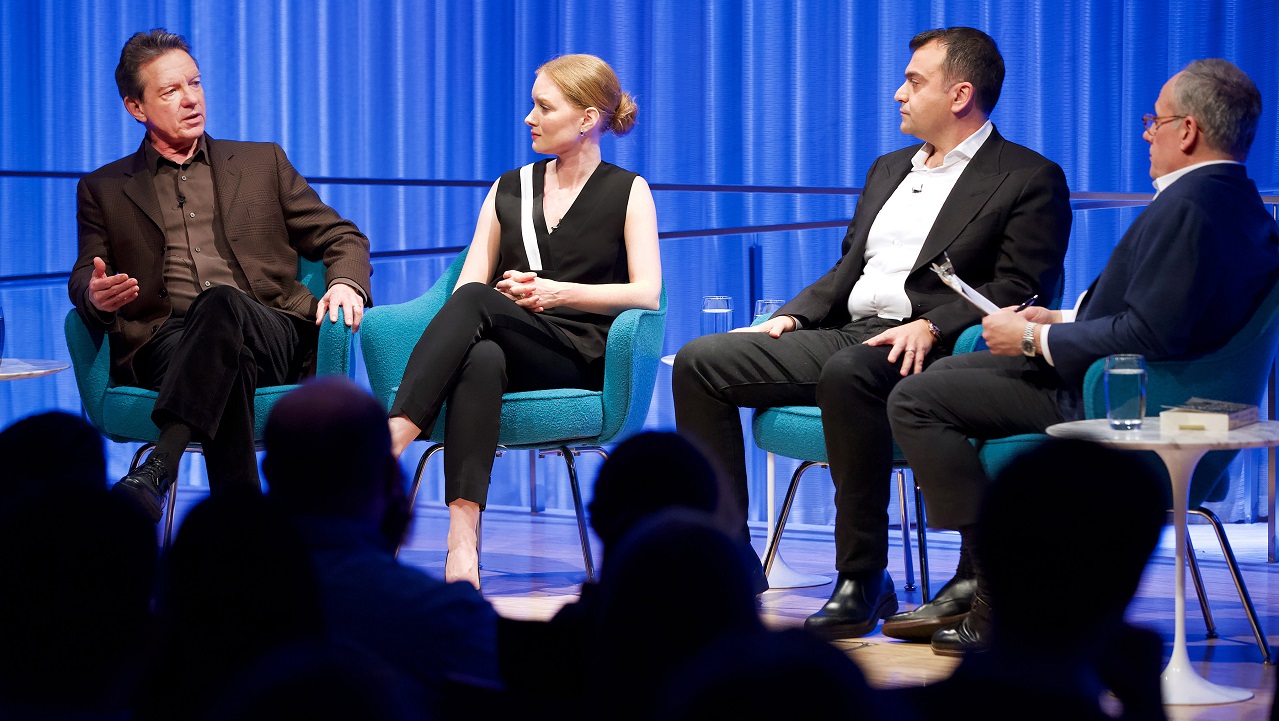 Three men and a woman take part in a moderated discussion on stage at the Museum. The woman is speaking, second from the left, as the three men listen. The black silhouettes of audience members are in the foreground.