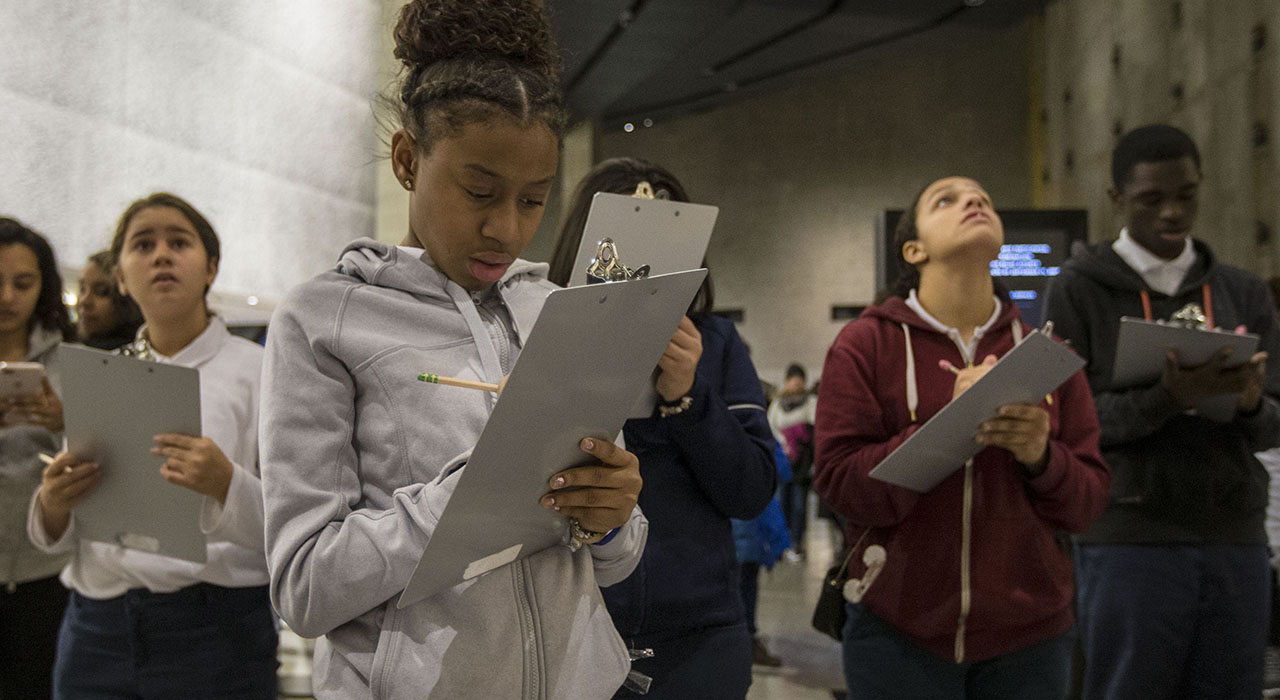 Group of middle school students in different skin tones write on clipboards inside the museum, with aluminum cladding visible in the background.