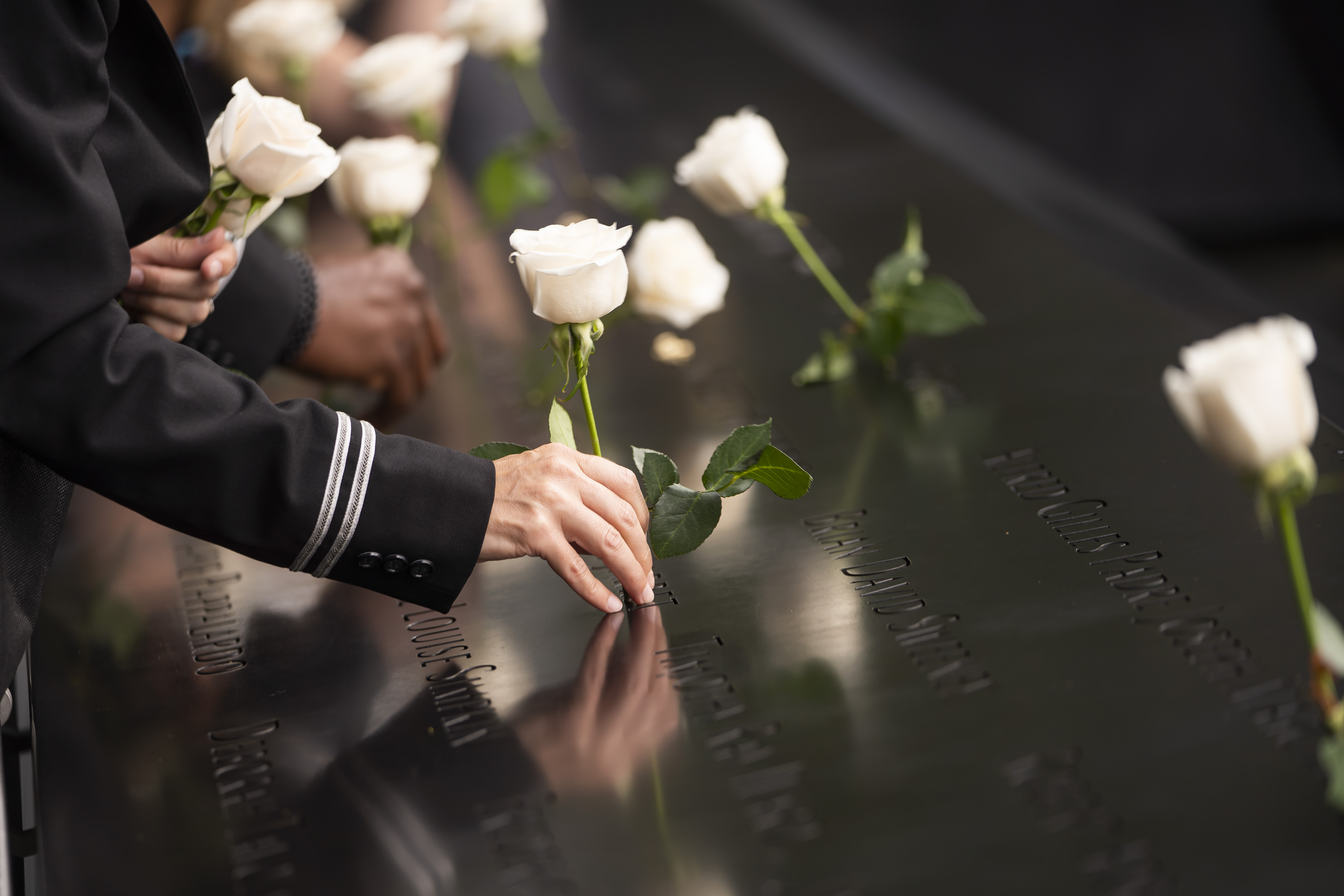 In this close up shot ot the 9/11 Memorial names parapet, a man's arm is shown from the elbow down as he places a white rose inside a victim's name on the Memorial. 