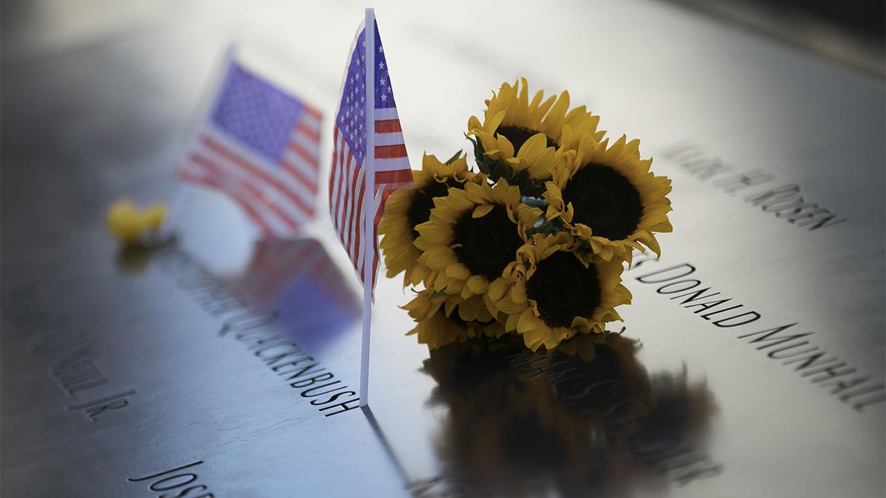 A bunch of sunflowers rest on the 9/11 Memorial, which is cast in a shimery yellow and blue light. Two American flags rest alongside it.