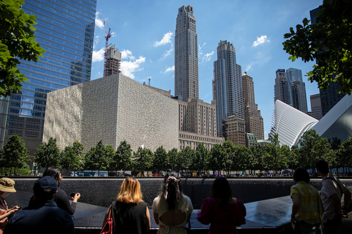 The Memorial lined with leafy trees, with visible visitors shown from the back and the lower Manhattan skyline