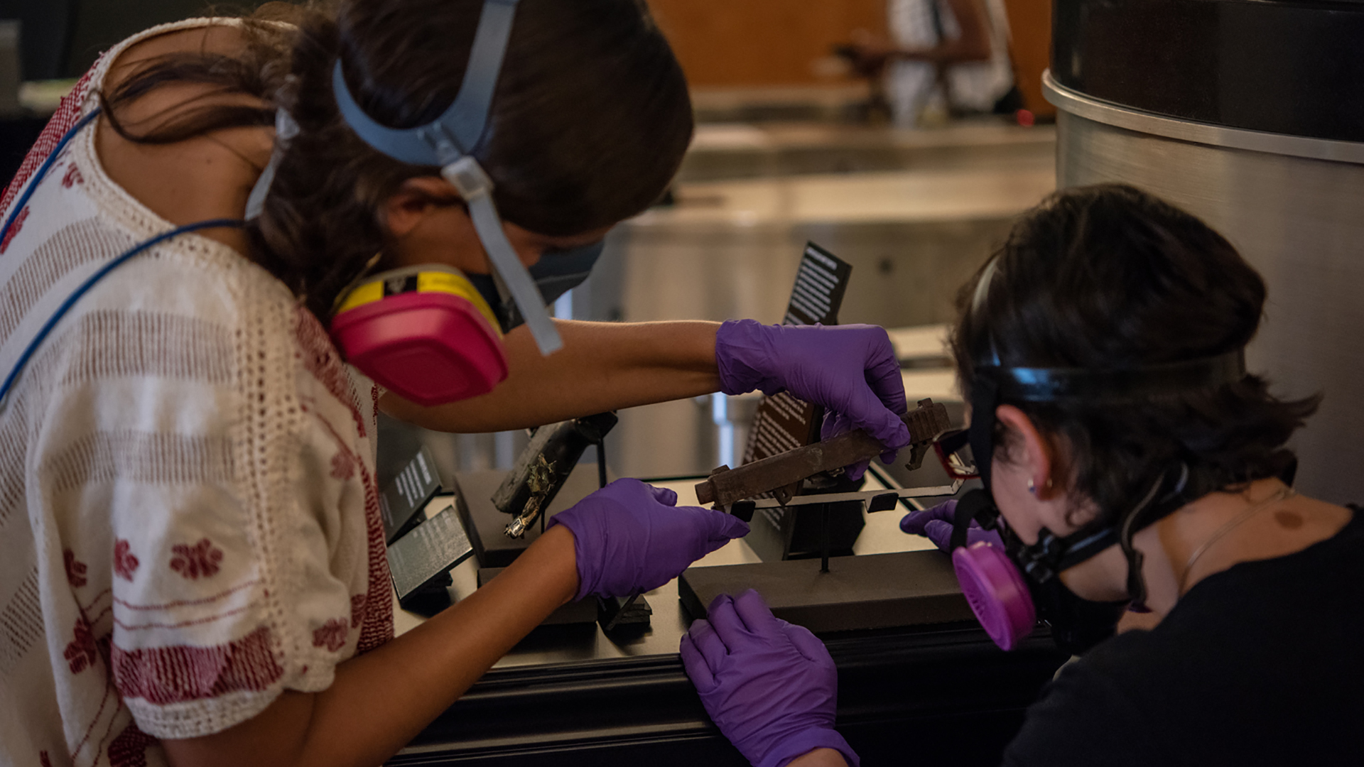 Two museum workers, in respiratory masks and surgical gloves, closely inspect a flat, rectangular artifact.