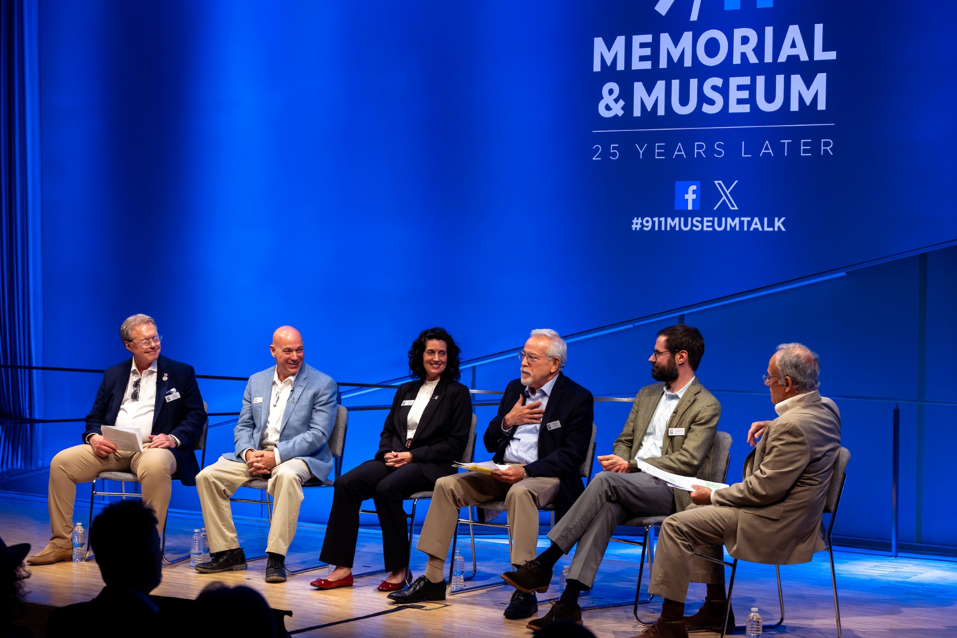 A group of six people sit on stage before a blue background with the 9/11 Memorial and Museum logo