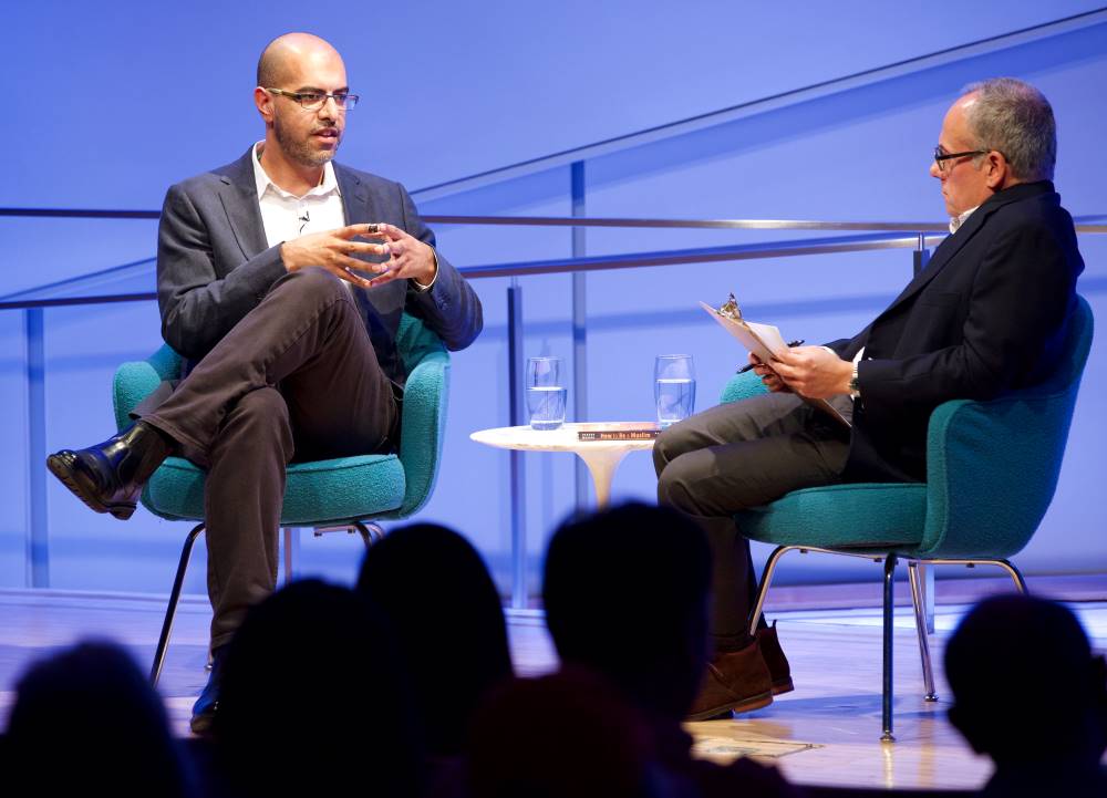 Author and news commentator Haroon Moghul presses his hands together as he speaks onstage at the Museum Auditorium. His legs are crossed and he is looking towards the audience. Clifford Chanin, the executive vice president and deputy director for museum programs, is seated next to him holding a clipboard. The heads of audience members are silhouetted in the foreground.