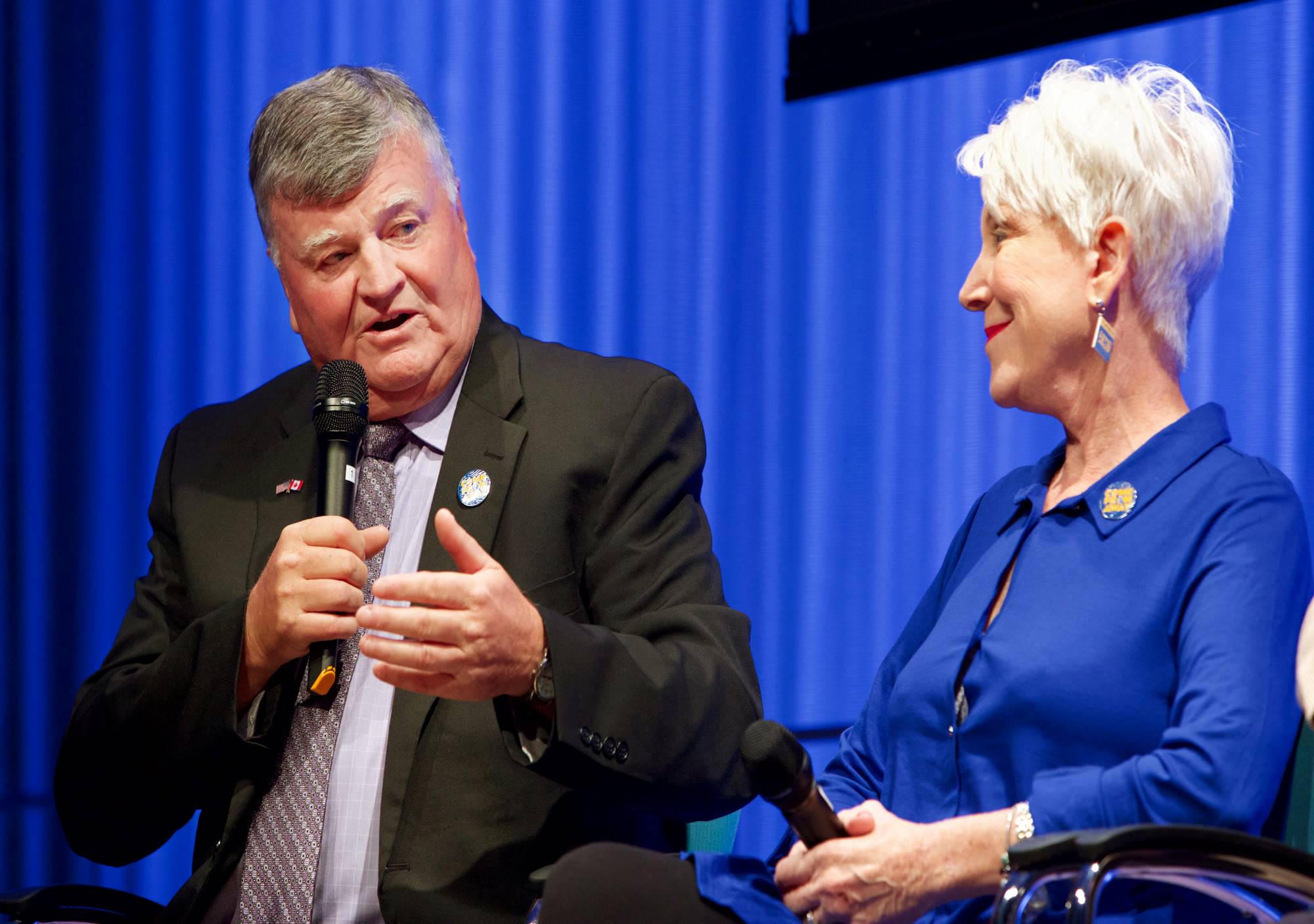 A man and woman behind the award-winning new musical “Come From Away” sit onstage during a public program in the Museum Auditorium in this close-up shot. The man has a microphone and is looking off to his left as a woman sitting next to him looks on.