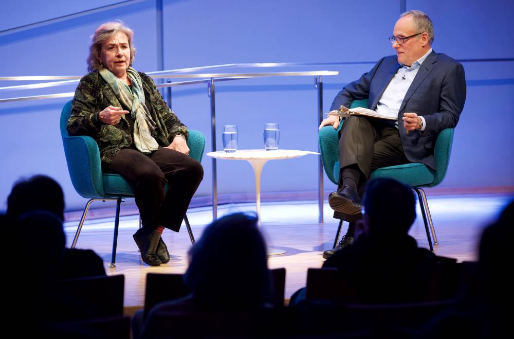 Dr. Sheila Carapico, professor of political science and international studies at the University of Richmond, looks towards the audience as she speaks onstage as part of the public program, Why Yemen Matters. Clifford Chanin, the executive vice president and deputy director for museum programs, is seated next to her and watching on as he holds a clipboard. Audience members in the foreground are silhouetted by the lights on stage.