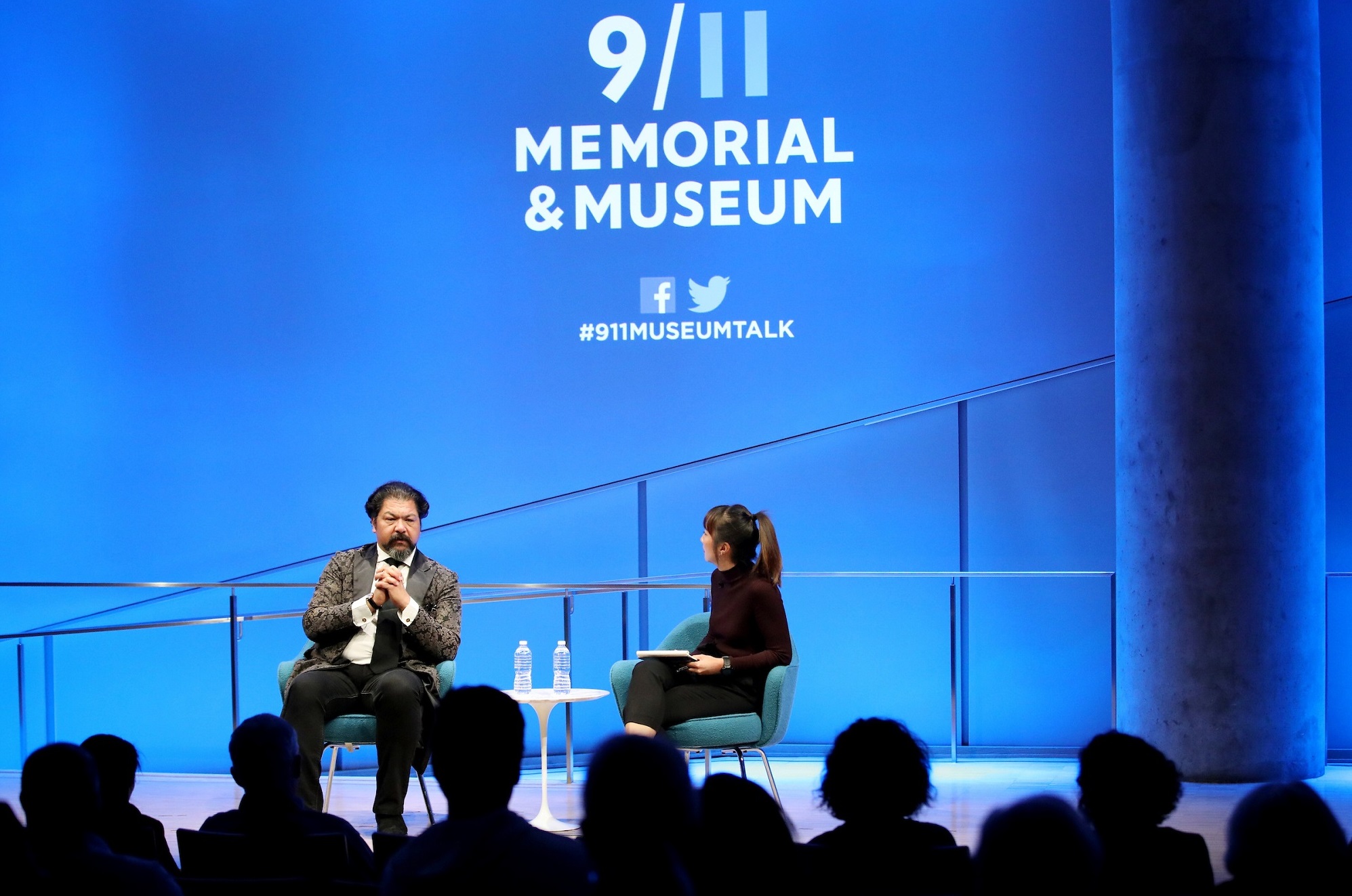 This wide-angle photo of the Museum Auditorium shows Cellist Karim Wasfi and a woman hosting the event seated onstage in the distance. Wasfi has his hands clasped together as he speaks. A wall behind the two participants is lit blue and features the logo of the 9/11 Memorial & Museum.