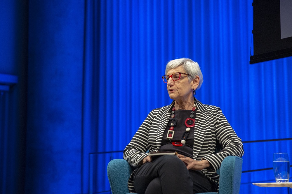 Harriet Senie, director of art history at the City College of New York, speaks onstage at the Museum Auditorium. She is seated in a chair in front of a curtain in the background that is illuminated blue.