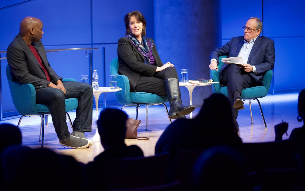 Three public program participants sit on a blue-lit auditorium stage. To the far left, a man in a gray blazer, red sweater, and jeans looks toward a woman speaking. The moderator, who sits with a clipboard in his lap, looks on. The heads of the audience members appear in the foreground in silhouette.